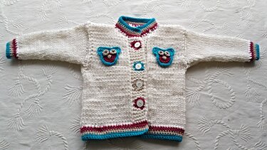 Cremefarbene, grob gestrickte Kinderjacke flach liegend mit mittiger Knopfleiste, türkis- und bordeauxfarbenen Kanten und zwei Eulen-Applikationen