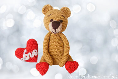 Gehäkelter brauner Amigurumi-Teddybär mit roten Herzfüßen und rotem Herzkissen mit 'Love'-Schriftzug.