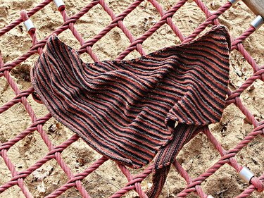 Gestricktes Dreieckstuch mit wellenförmigen Streifen in Rot, Dunkelgrau und Hellgrau auf einem roten Seilnetz über Sand