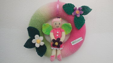 Handgestrickter Willkommenskranz in Grün und Pink mit gestrickter Elfenfigur, Blumen und Blättern, an einer weißen Wand hängend.