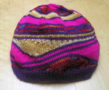 Gestrickte Damenmütze in Pink, Violett und Gold mit Rippenbund auf hellem Holzuntergrund