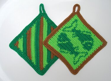 Zwei gehäkelte Topflappen, einer gestreift in Grün und Braun, der andere mit Blättermuster in Grün und braunem Rand, auf weißem Hintergrund liegend.