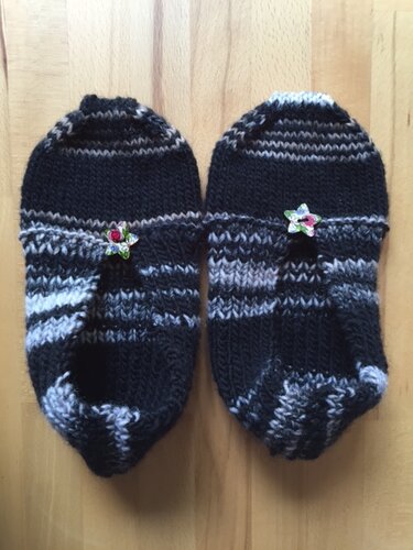 Paar schwarz-grauer gestrickter Hausschuhe mit Blumenknöpfen auf hellem Holz
