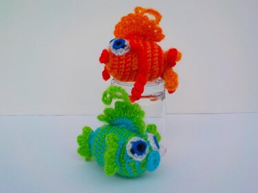 Zwei gehäkelte Amigurumi-Fische, orange auf einem Glas und grün-blau davor