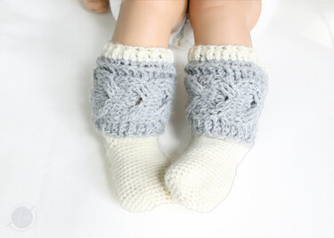 Nahaufnahme von Babybeinen mit cremefarbenen Socken und grauen, zopfmustergehäkelten Stulpen.