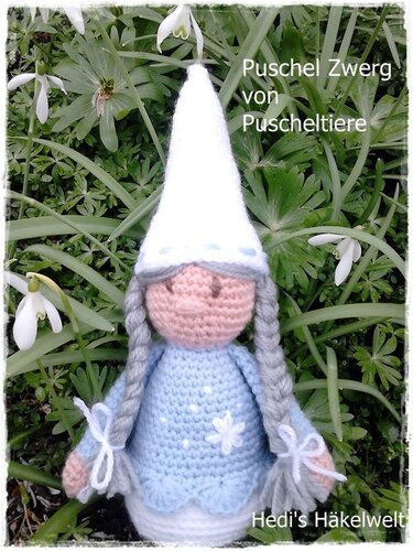 Gehäkelte Zwergenpuppe mit weißer Zipfelmütze, grauen Zöpfen und blauem Kleid zwischen grünen Pflanzen und Schneeglöckchen