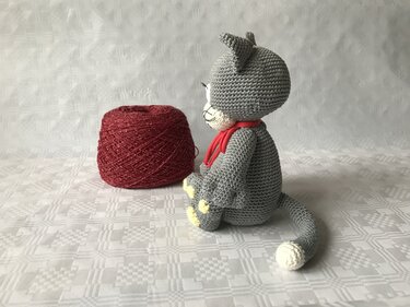 Gehäkelte graue Amigurumi-Katze mit rotem Schal neben roter Garnrolle, seitliche Ansicht
