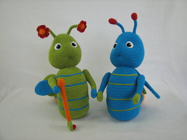 Zwei gehäkelte Amigurumi-Insekten: eine grüne mit Blüten an den Fühlern und orangefarbenem Stab, eine blaue mit roten Fühlerspitzen, vor hellem Hintergrund.