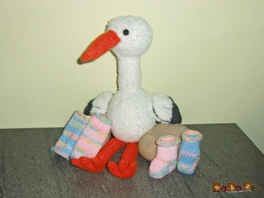 Gestrickter weißer Amigurumi-Storch mit orangefarbenem Schnabel und roten Beinen zwischen pastellgestreiften Babysocken