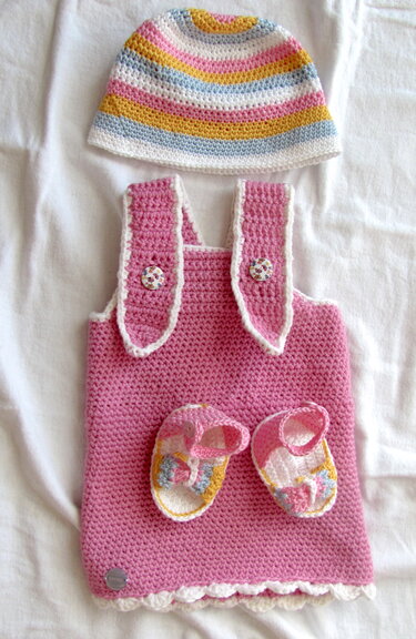 Gehäkeltes rosa Baby-Trägerkleid mit gestreifter Mütze und Schühchen auf weißem Stoff