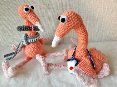 Zwei gehäkelte rosa‑apricot Amigurumi‑Vögel mit langen Hälsen, schlanken Beinen, Schals und kleiner Tasche