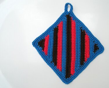 Gehäkelter, rautenförmiger Topflappen mit blauem Rand und rot-dunkelbraunen Streifen, mit Aufhängeschlaufe.