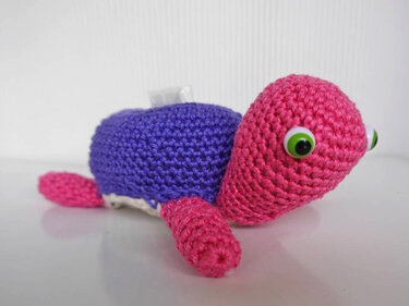 Gehäkelte Amigurumi‑Schildkröte mit rosa Kopf und Beinen, lila Panzer und großen grünen Augen auf hellem Untergrund.