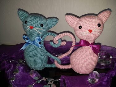 Zwei gehäkelte Amigurumi-Katzen in Türkis und Rosa, deren Schwänze ein Herz formen, auf violettem Stoff dekoriert.