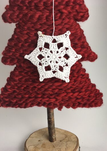 Weiße gehäkelte Schneeflocke an rotem, grob gestricktem Deko‑Tannenbaum mit Holzstamm
