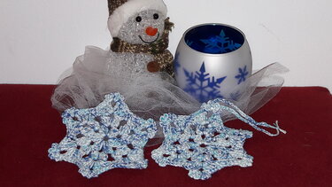Zwei gehäkelte blau-weiße Schneeflocken vor Schneemann und Teelichtglas auf rotem Untergrund.