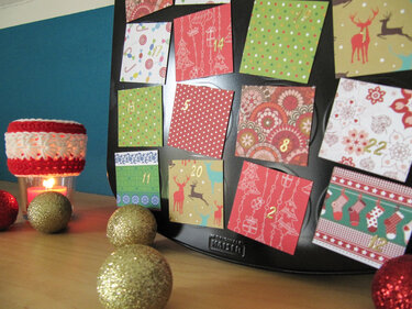 Adventskalender aus schwarzem Muffinblech mit nummerierten, gemusterten Papierklappen; daneben Kerze in gehäkelter Hülle und glitzernde Christbaumkugeln.