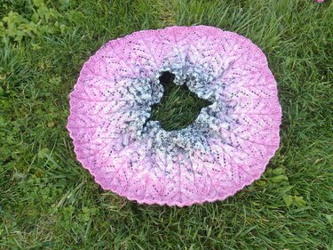 Nahaufnahme eines runden gestrickten Loops mit Ajourmuster und Farbverlauf von rosa zu grau auf grünem Rasen
