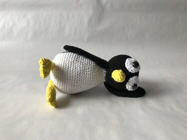 Gehäkelter Amigurumi‑Pinguin liegend, schwarzer Kopf, weißer Körper, gelber Schnabel und Füße