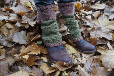 Zweifarbige, gestrickte Beinstulpen in Dunkelrot und Moosgrün über dunklen Schuhen auf trockenem Herbstlaub.