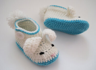 Weiß-blaue gehäkelte Babyschuhe mit Hasenohren, gestickten Augen, rosa Nase, Bommel und Knopfverschluss.