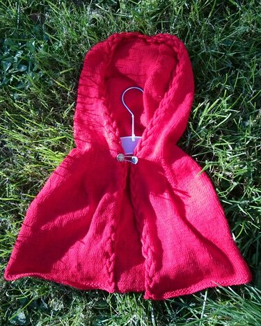 Märchenhaftes rotes gestricktes Kapuzencape mit gezopfter Kante und dekorativem Verschluss auf Kleiderbügel im Gras
