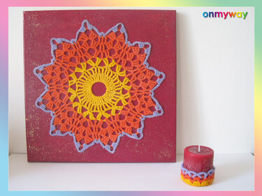 Rote Leinwand mit gehäkeltem Mandala in Gelb, Orange und Lila; rote Kerze mit Häkelüberzug.