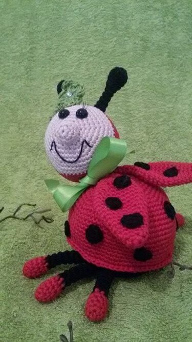 Gehäkelter Marienkäfer-Amigurumi in Rot und Schwarz mit rosa Kopf, grüner Schleife und kleiner Blüte vor grünem Hintergrund.
