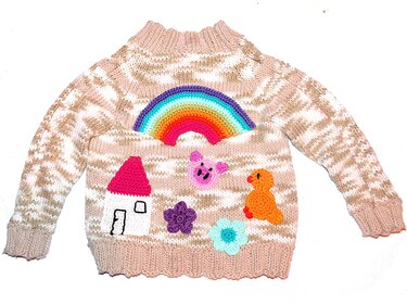 Beige-weißer gestrickter Kinderpullover mit bunten Applikationen: Regenbogen, Haus, Schwein, Ente und Blumen
