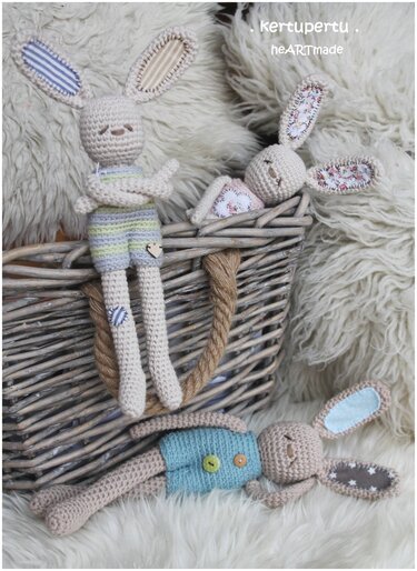 Drei gehäkelte Amigurumi-Hasen mit unterschiedlicher Kleidung auf hellem Fellteppich neben einem Weidenkorb.