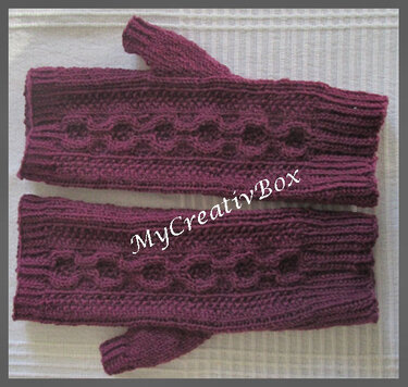 Zwei violett-bordeauxfarbene gestrickte Armstulpen mit Zopfmuster und Daumenloch auf hellem Untergrund