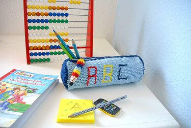Hellblaues gehäkeltes Federmäppchen mit bunten Buchstaben ABC und gehäkeltem Bleistift-Anhänger auf Schreibtisch neben Rechenrahmen und Stiften