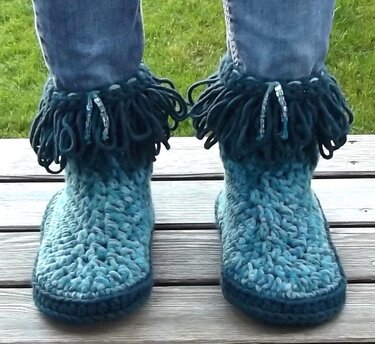 Paar gehäkelter türkiser Damenstiefel mit Fransen am Schaft, getragen zu Jeans auf Holzplanken.