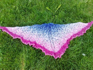 Gestricktes Dreieckstuch mit blau-rosa Farbverlauf, Ajourmuster und gezackter Spitzenborte auf Rasen ausgelegt