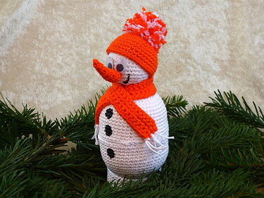 Gehäkelter Amigurumi-Schneemann mit orangefarbener Mütze und Schal auf Tannenzweigen