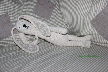 Cremefarbener gehäkelter Amigurumi-Hase liegt seitlich auf gestreifter Bettwäsche, lange Ohren mit weißer Stickerei sichtbar.