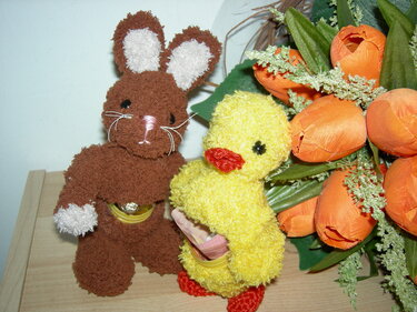 Brauner gehäkelter Hase und gelbes gehäkeltes Küken neben orangefarbenen Tulpen auf einer Holzoberfläche.