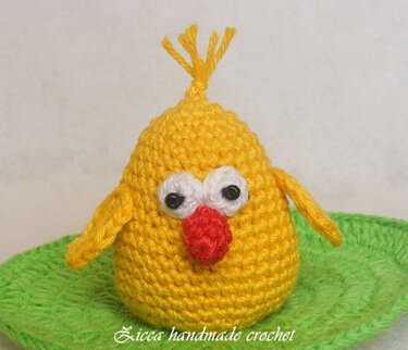 Handmade yellow crochet chick with white eye patches, red beak and top tuft on a green crocheted base