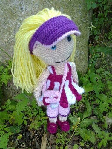 Gehäkelte Amigurumi-Puppe mit gelbem Garnhaar, violetter Schirmmütze und rosa-weiß gestreiftem Outfit im Gras