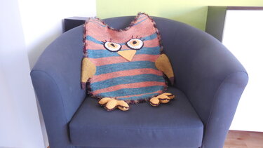 Gestreiftes Eulen-Kissen mit applizierten Augen, Schnabel und gelben Flügeln auf einem dunklen Sessel