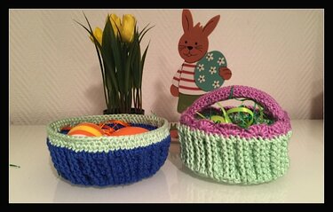 Zwei gehäkelte Körbe: flache blau-hellgrüne Schale mit Dekoeiern und kleines hellgrün-lila Körbchen mit Henkel und Ostergras; gelbe Tulpen und Holzosterhase im Hintergrund.