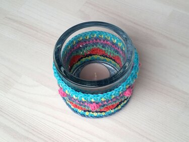 Glas-Teelichthalter mit gehäkeltem Überzug in Türkis, Pink und Orange auf hellem Holztisch, Teelicht im Inneren.