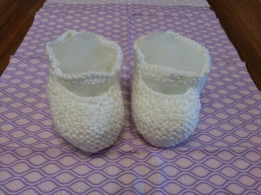 Paar weißer handgestrickter Babyschuhe mit Riemchen auf lila Stoff
