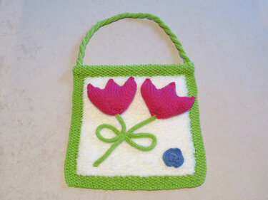 Gestricktes Quadrat mit zwei pinken Tulpen, grünem Rand, Aufhängeschlaufe und blauem Punkt
