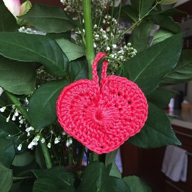 Rotes gehäkeltes Herz mit Schlaufe zwischen grünen Blättern und weißen Blüten