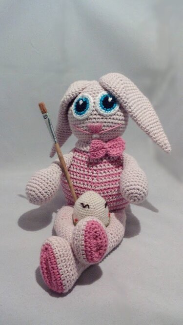 Gehäkelter rosa-weißer Amigurumi-Hase mit blauen Augen und rosa Fliege, der einen Pinsel hält; ein kleines gehäkeltes Osterei sitzt vor ihm.