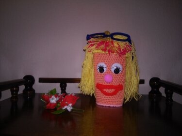 Gehäkelte Amigurumi-Figur mit gelbem Garnhaar, großen Augen, rosa Nase, rotem Mund und blauer Brille auf dunklem Holztisch.