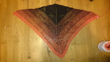 Vogelperspektive: gehäkeltes Dreieckstuch mit Farbverlauf auf Holzboden, Garnknäuel und Maschenmarkierer am Rand