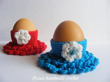Zwei gehäkelte Eierwärmer (rot und türkis) mit weißen Blüten, jeweils mit braunem Ei