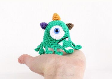 Palm-sized green crocheted one-eyed monster egg cosy with purple, orange and brown horns, white eye and fringe base, sitting on a hand